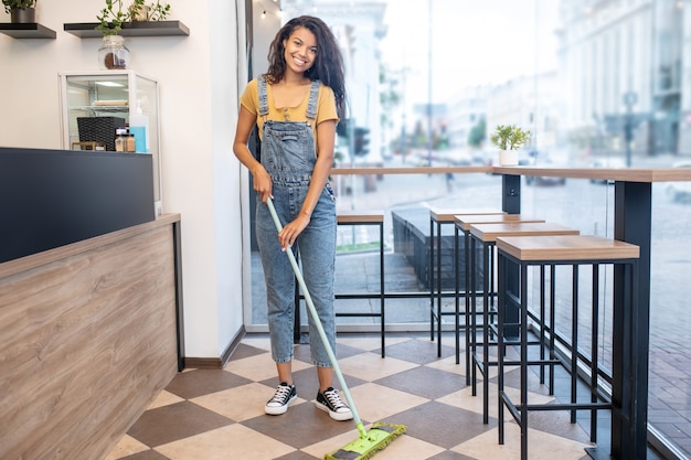 Work, cafe. Happy woman with long dark hair with mop standing near bar in cafe doing wet cleaning