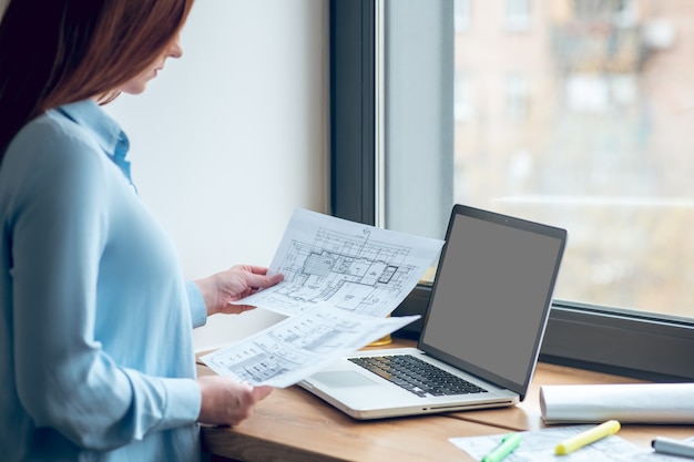 Foto lavoro, concentrazione. donna concentrata dai capelli lunghi in camicetta leggera che guarda i piani di costruzione su carta in piedi vicino alla finestra all'interno durante il giorno