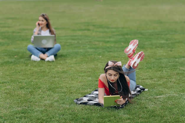 Work on freelance. Relax and be happy. Student learning. Two pretty girls friends sitting on the green grass.