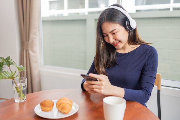 自宅のコンセプトから仕事をするヘッドフォンを身に着けている若い女性が、デザートとテーブルの上で飲み物を飲みながらお気に入りのプレイリストを聴いている瞬間を楽しんでいます。