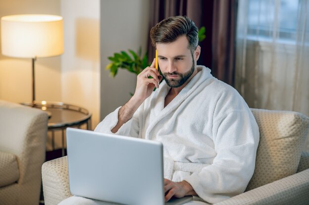 Work from home. Young bearded man in a white bath robe working on a laptop and talking on the phone