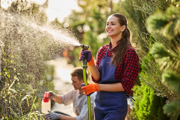 Work in the garden. Smiling girl gardener sprays water and a guy sprays fertilizer on plants in the beautiful nursery-garden on a sunny day. .