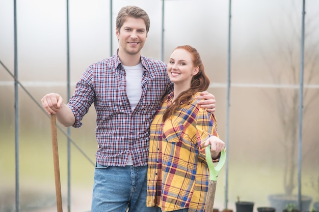 Work, hobby. Smiling young adult woman and man in plaid shirts with garden tools standing in their greenhouse