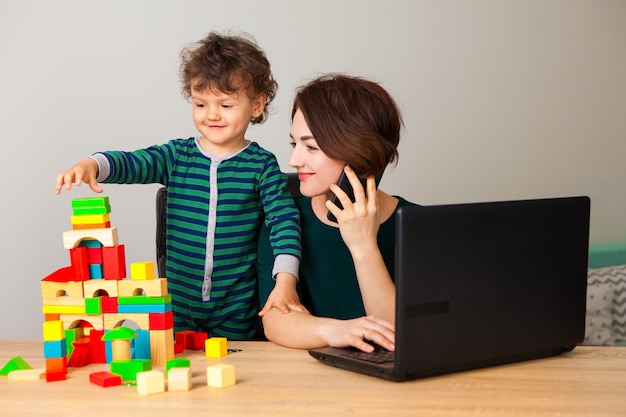 Work at home. A woman sits working on a laptop and talking on the phone, looking at the child as he plays cubes and builds a large multi-story house.