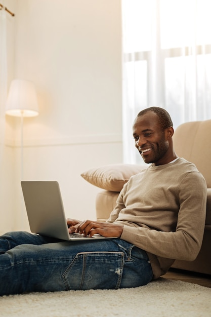 Work. Joyful bearded afro-american man smiling and working on the laptop and typing while sitting on the floor near the couch and a lamp in the background