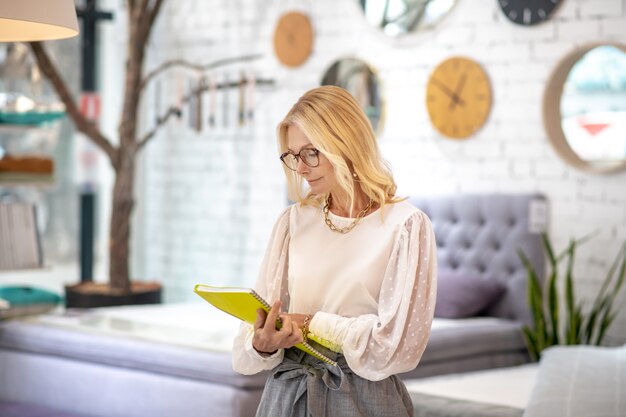 Work moment. Blonde woman with glasses standing in the showroom with a large yellow notebook in her hands, looking at her with interest.