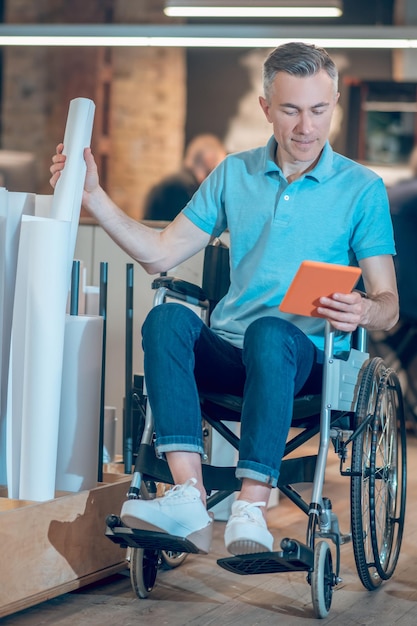 Work moment. Man on wheelchair looking closely at tablet holding roll of paper near special stand in office space