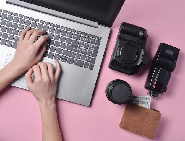The work of the photographer, photo retouching. Photographic equipment, female hands typing on laptop keyboard on pink pastel background, top view, concept of freelancing, flat lay