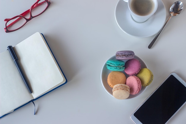 Work table with writing pad, glasses, coffee cup, mobile, and plate full of colorful macaroons, on light background.