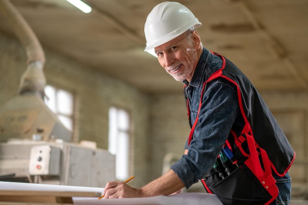 Work time. Mature man in protective helmet working in a workshop and looking contented