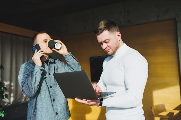 Work of two male colleagues standing in office together while exchanging ideas with laptop in meeting room and analyzing work schemes Work in a modern spacious office