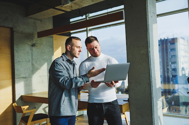 Work of two male colleagues standing in office together while exchanging ideas with laptop in meeting room and analyzing work schemes Work in a modern spacious office