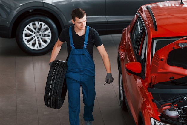 Worker in black and blue uniform holds car wheel and walks with in near red automobile