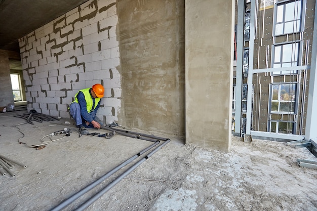 작업자 또는 건축업자가 보호 복 및 안전모를 착용하고 건설중인 건물의 평면에 현대적인 도구를 사용하여 플라스틱 파이프를 설치하고 있습니다.