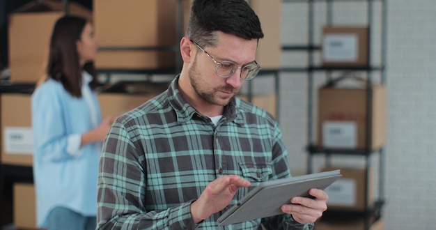 Worker checking inventory writing notes on tablet A man works in a warehouse with rows of shelves with boxes packages