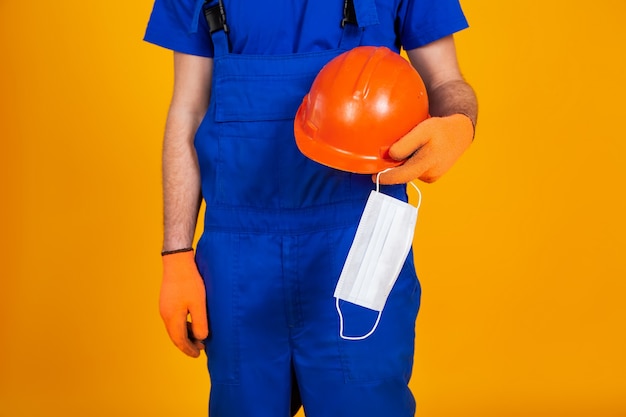 Worker in construction overalls and gloves on a yellow background, holds a protective helmet and a medical mask in his hand. the concept of the economic crisis, unemployment production, coronavirus