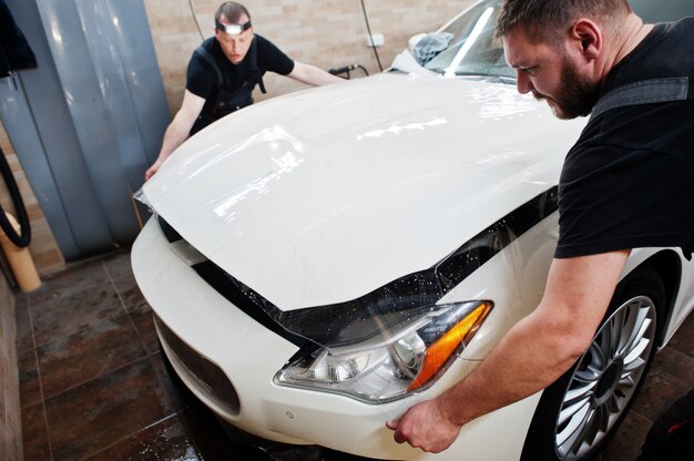 Worker in detailing garage put polyurethane anti-gravel film cover in white luxury car.