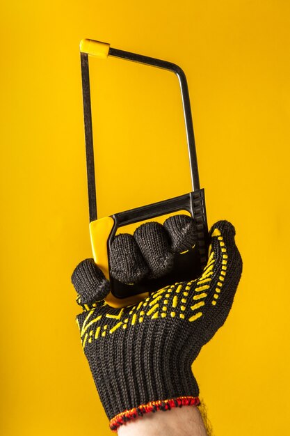 手袋をはめた労働者の手は、黄色の背景にのこぎりまたは弓のこを保持します。建物や改修のアイデア
