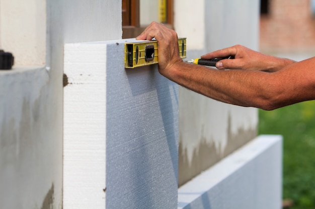 Worker hand with level and knife measuring and cutting white rigid polyurethane foam sheet on wall at newly installed plastic window. Modern technology, renovation, insulation concept.