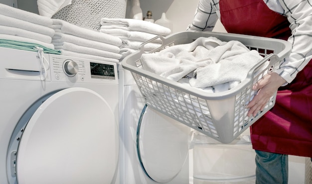 Worker in the hotel holds a lot of towels in hands near dry\
machine in the laundry