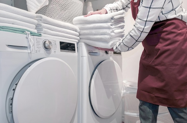 Worker in the hotel holds a lot of towels in hands near Dry machine in the laundry