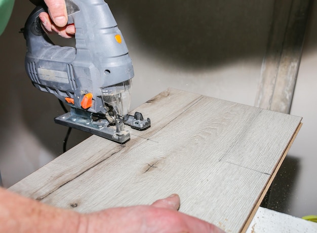 Worker is holding in hands power jigsaw and cutting laminate\
flooring in apartment maintenance repair works renovation with\
scroll saw wooden parquet floor planks indoors