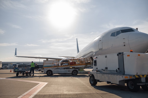 Worker loading baggage on conveyor belt to an airplane outdoors on a daytime. Plane, shipping, transportation concept