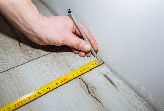 Worker making laminate flooring in apartment measure tape and\
pencil in hands maintenance repair renovation wooden parquet planks\
indoors