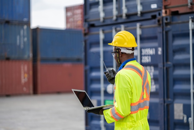 Рабочий в защитном комбинезоне безопасности с желтой каской и использует контейнер для проверки рации на грузовом складе. транспорт импорт, экспорт логистика промышленные услуги