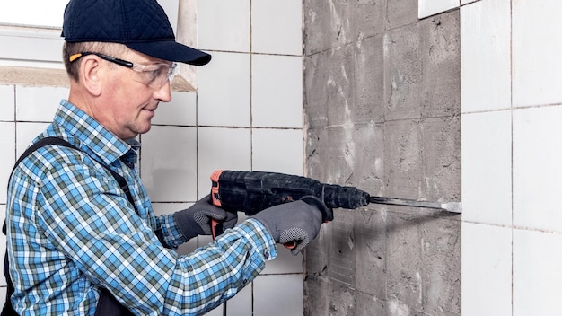 A worker in overalls and protective glasses dismantles facing tiles with a perforator