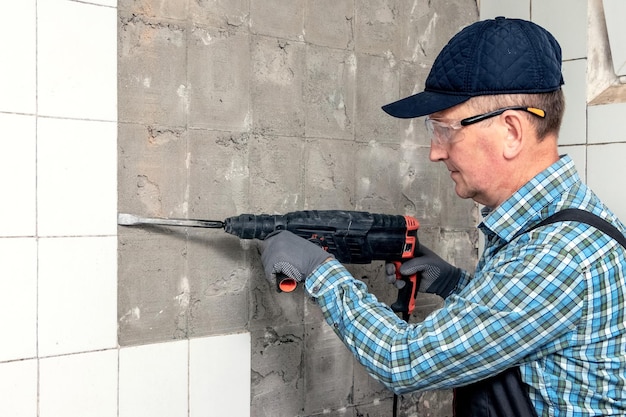 A worker in overalls and protective glasses dismantles facing tiles with a perforator