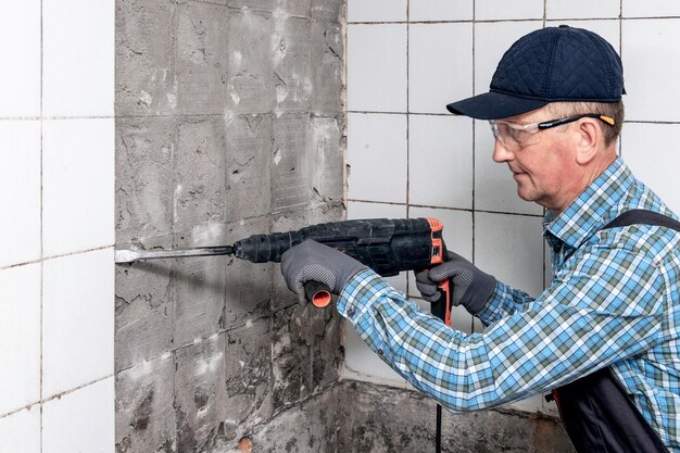 A worker in overalls and protective glasses dismantles facing tiles with a perforator