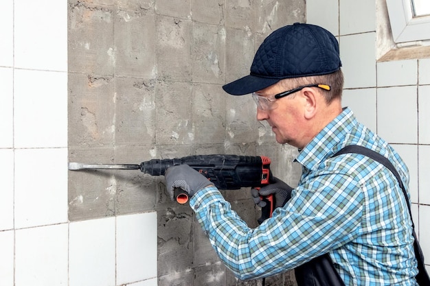 A worker in overalls and protective glasses dismantles facing tiles with a perforator