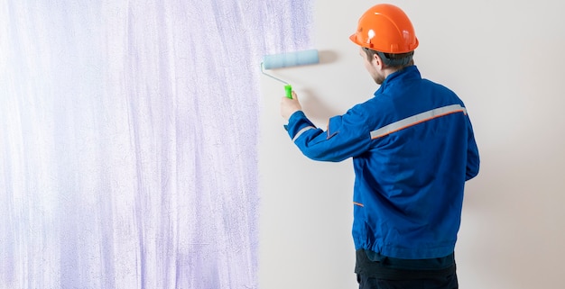 Художник-рабочий красит поверхность стены валиком, мужчина за работой украшает комнату
