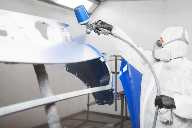 Worker paints bumper of auto with spray gun White car in a paint chamber during repair work