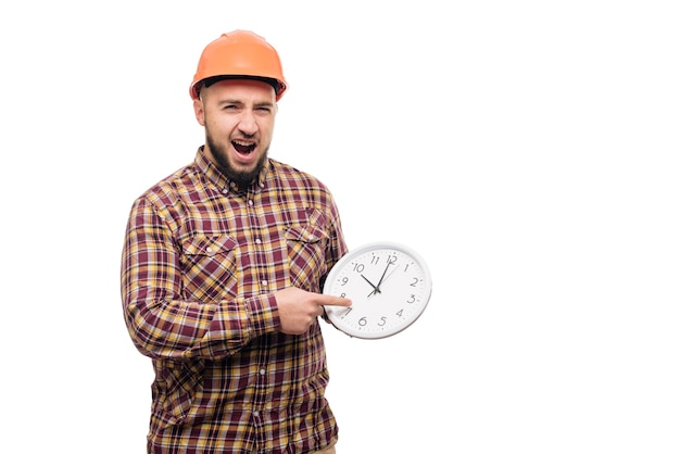 白の目覚まし時計を保持している保護ヘルメットの労働者。働く時間。建物の建設時間