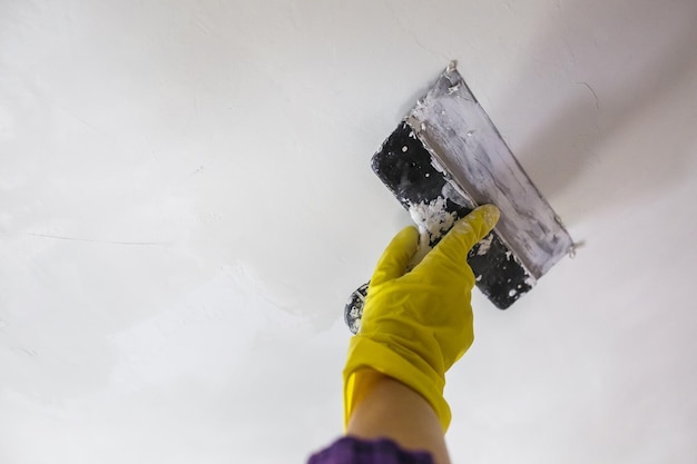 Worker's hand in yellow gloves holding putty knife patching a hole with spatula with plaster or putty in white wall Renovation and repair process remodeling interior of room at apartment building