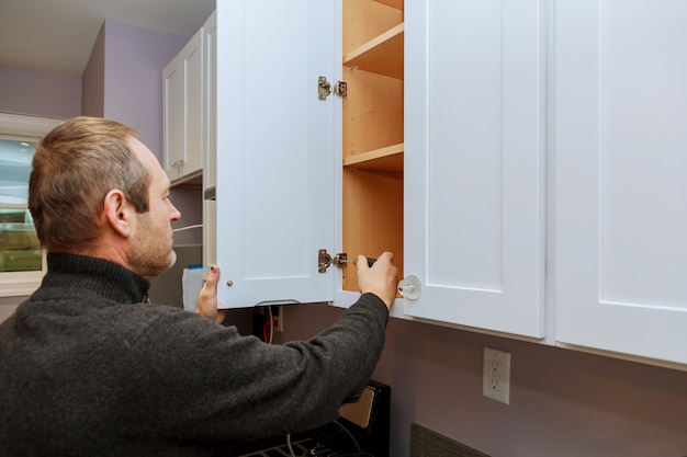 Photo worker sets a new handle on the white cabinet with a screwdriver installing kitchen cabinets