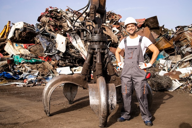 worker standing by hydraulic industrial machine used for lifting scrap metal parts in junk yard