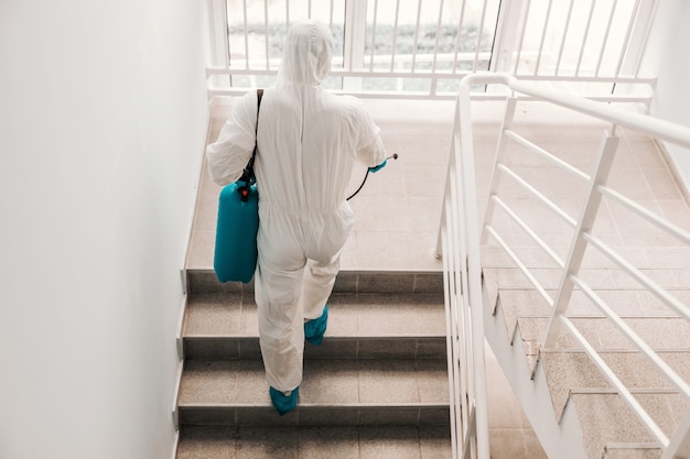 Worker in sterile uniform, with gloves and facial mask sterilizing stairs in school.