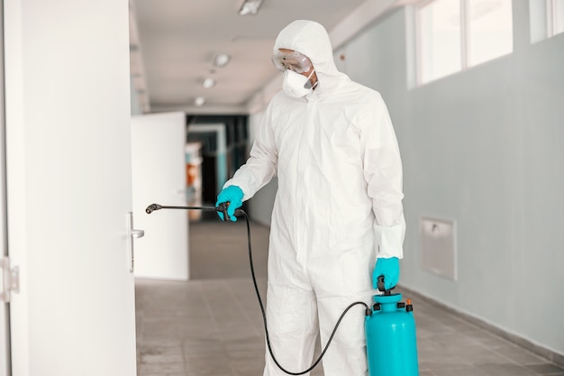 Worker in sterile white uniform, with mask and glasses holding sprayer with disinfectant and spraying door in school.