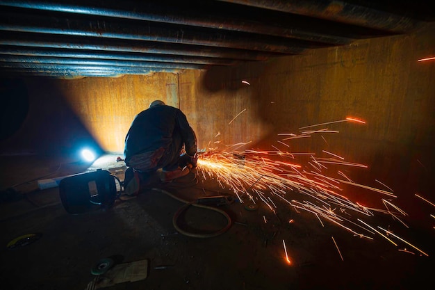 Worker using electric wheel spark grinding metal is part of machinery plate tank beam pipe construction flash spark inside confined