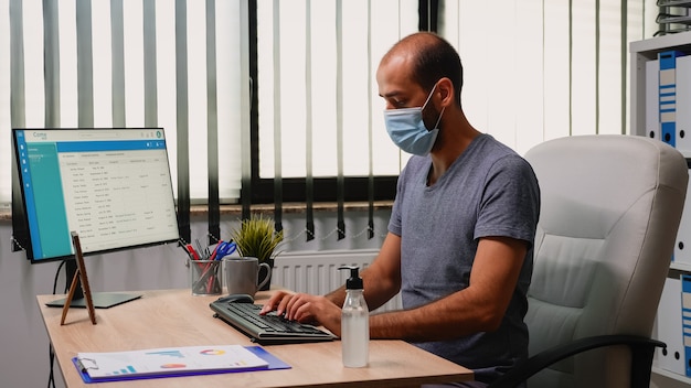 Lavoratore che indossa una maschera di protezione con gel igienizzante durante il coronavirus. imprenditore che lavora in un nuovo normale posto di lavoro in azienda che pulisce le mani utilizzando alcol antibatterico contro il virus corona.