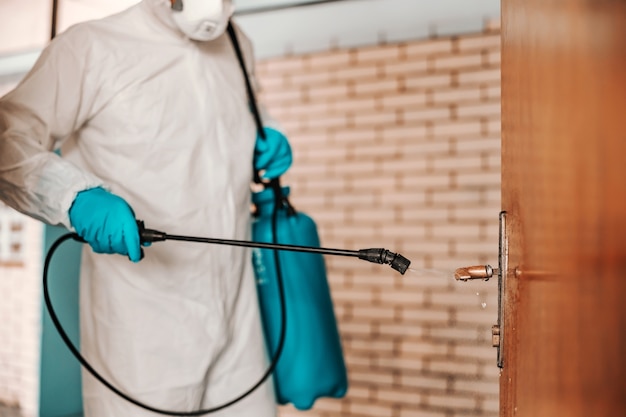 Photo worker in white sterile uniform, with rubber gloves and mask on holding sprayer with disinfectant and sterilizing doors in school.