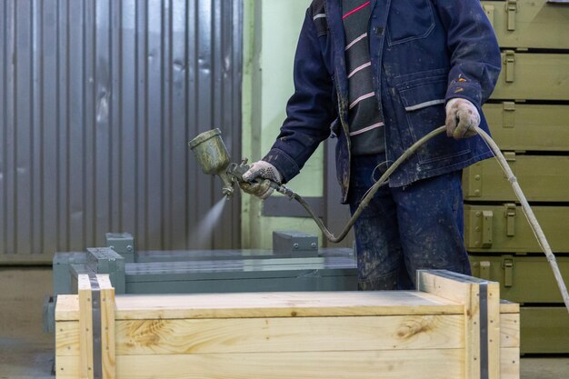 Worker with airbrush painting big wooden crates to gray color