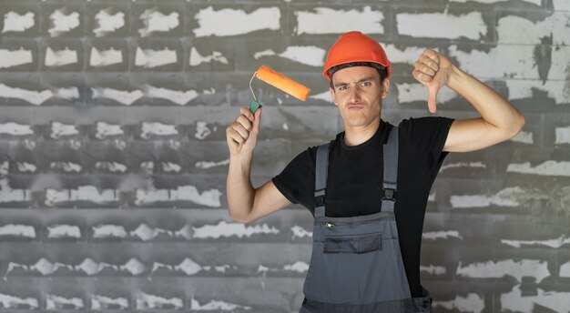 Worker with orange helmet helmet near a wall of stones. holding a roller in his hands thumb down