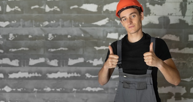 Worker with orange helmet helmet near a wall of stones. The thumb of both hands is raised