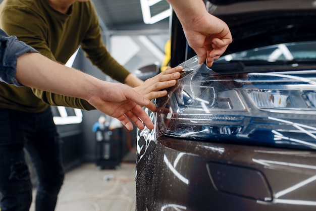 Рабочие наносят автомобильную защитную пленку на переднее крыло. Установка покрытия, защищающего краску автомобиля от царапин. Новый автомобиль в гараже, процедура тюнинга