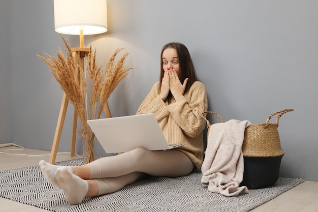 Рабочие онлайн-задачи Телеработа на дому Шокированная женщина с коричневыми волосами в свитере, работающая на компьютере, имеет проблемы с проектом, покрывающим рот руками, сидя на полу в домашнем интерьере