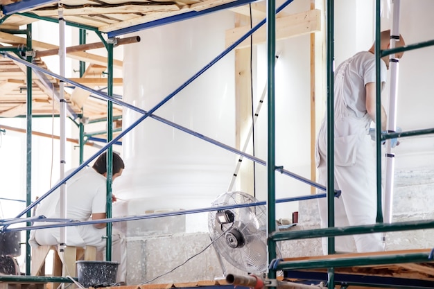 Workers restore white walls on scaffolding in large room